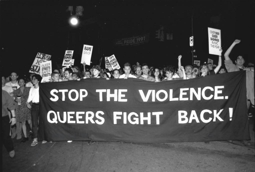 black and white photo of large group of protestors carrying a banner that reads "Stop the Violence. Queers Fight Back!"