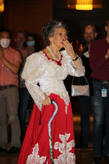 older Asian woman in sequined red and white dress. She is wearing pearls and holding a cane. She is standing in the center of a room while those around her applaud.