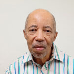 headshot portrait of older Black man in a striped button-up shirt, standing before a gray background