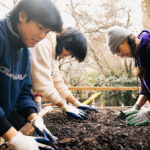 Three people wearing garden gloves sifting dirt on a flat screen sifter.