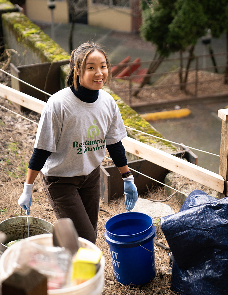 A woman wearing a grey t-shirt that says "Restaurant 2 Garden" and garden gloves carries a bucket through a community garden.