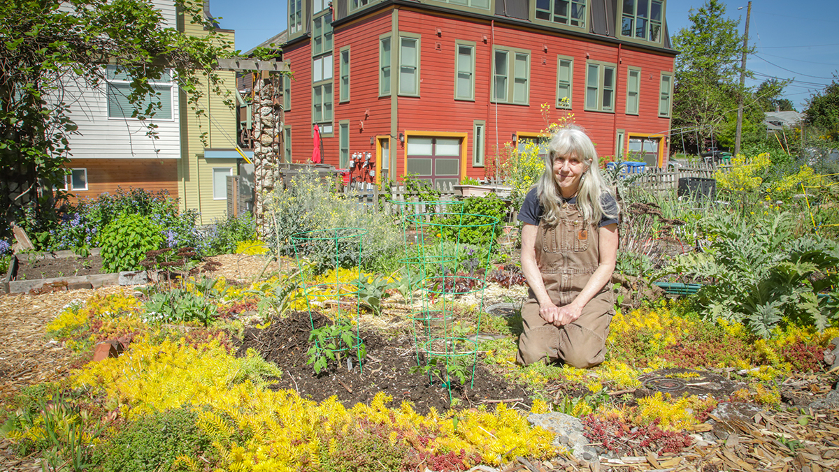 An older woman with long gray hair and tan coveralls kneels in a community garden bed with apartment buildings visible in the background
