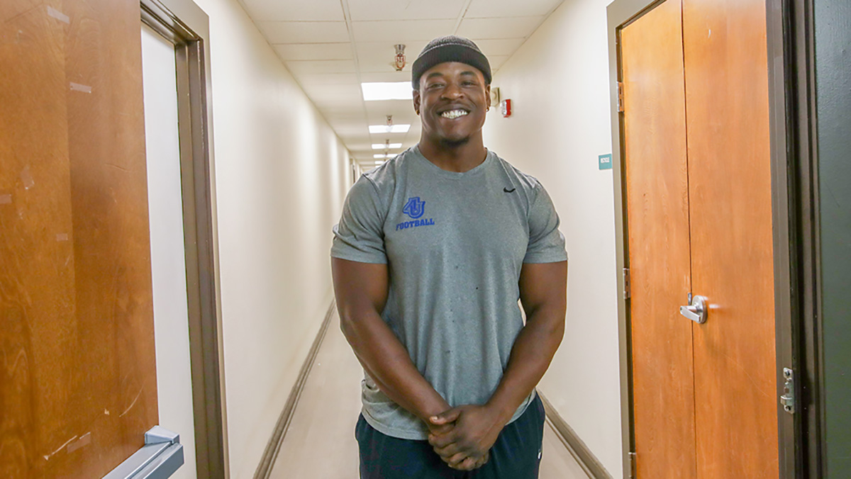 A Black man wearing a gray t-shirt and black beanie smiles while standing in a hallway