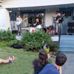 A group of people sit on a residential front lawn watching a band perform on a porch.
