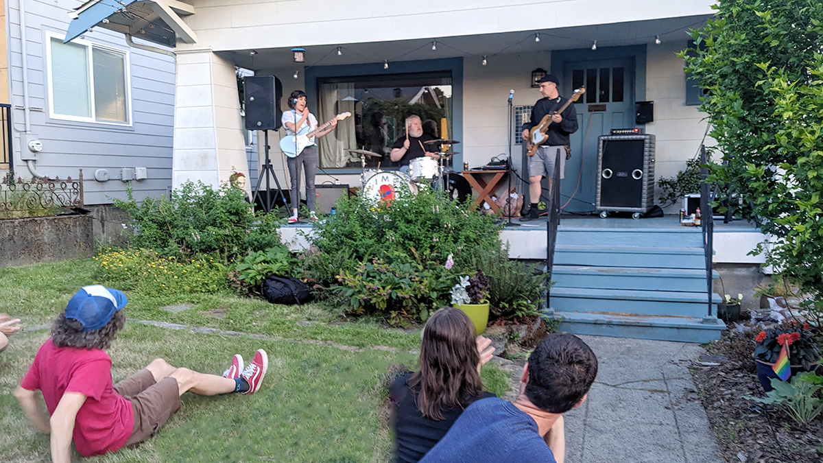 A group of people sit on a residential front lawn watching a band perform on a porch.