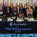 group of Indigenous and urban Native leaders standing with City of Seattle leaders behind a table. The table is draped with a black banner that reads "City of Seattle Tribal Nations Summit."