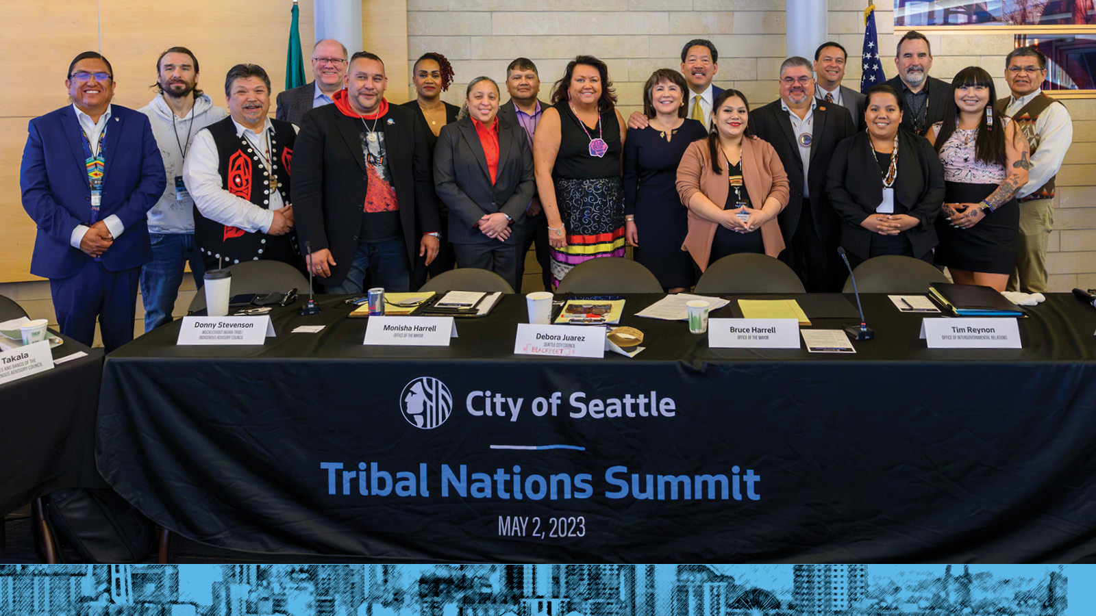group of Indigenous and urban Native leaders standing with City of Seattle leaders behind a table. The table is draped with a black banner that reads 