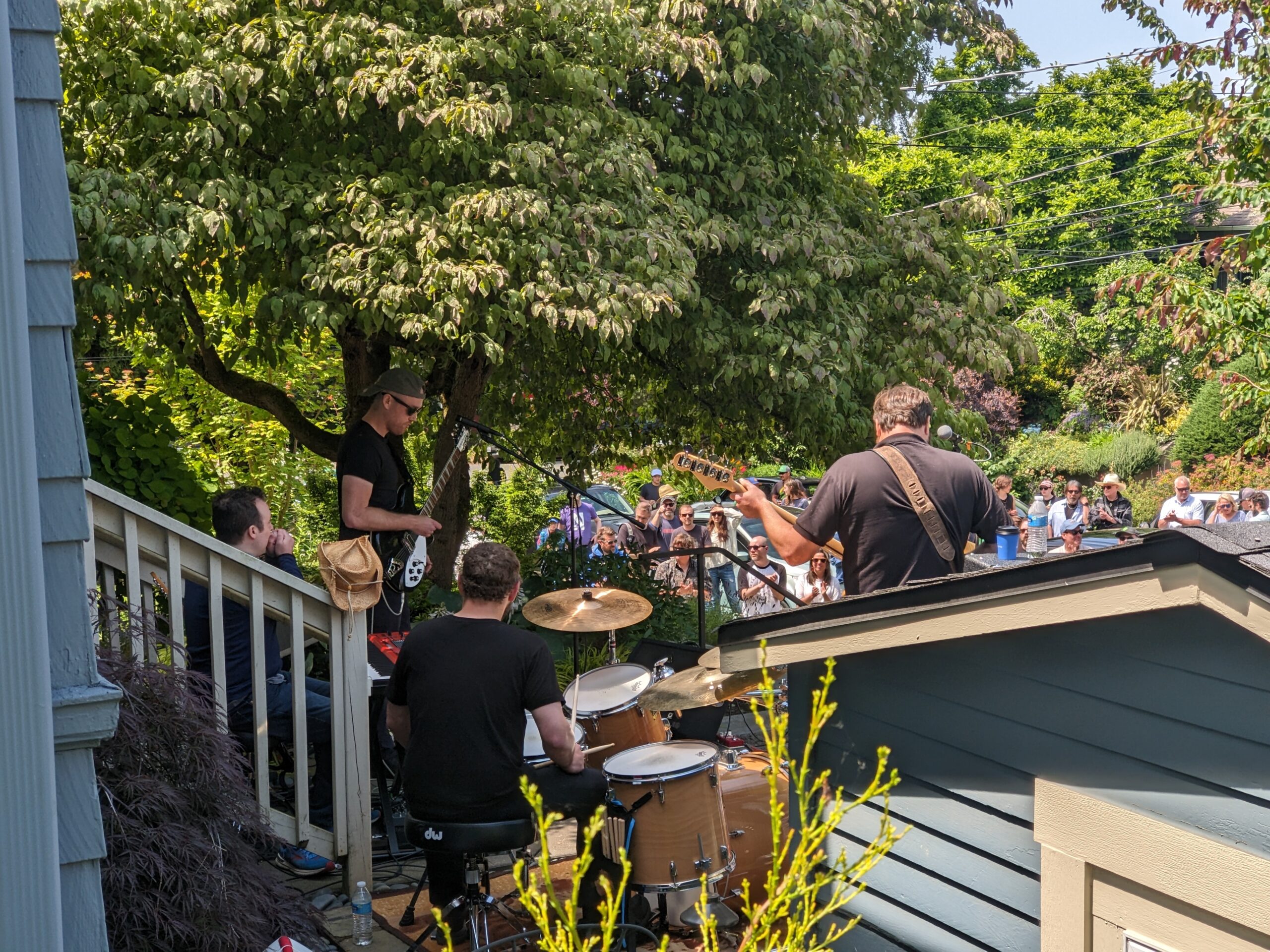 Shot of a band with a guitar, bass, keyboards, and drums playing on a residential front porch with people standing on the sidewalk watching in the background.