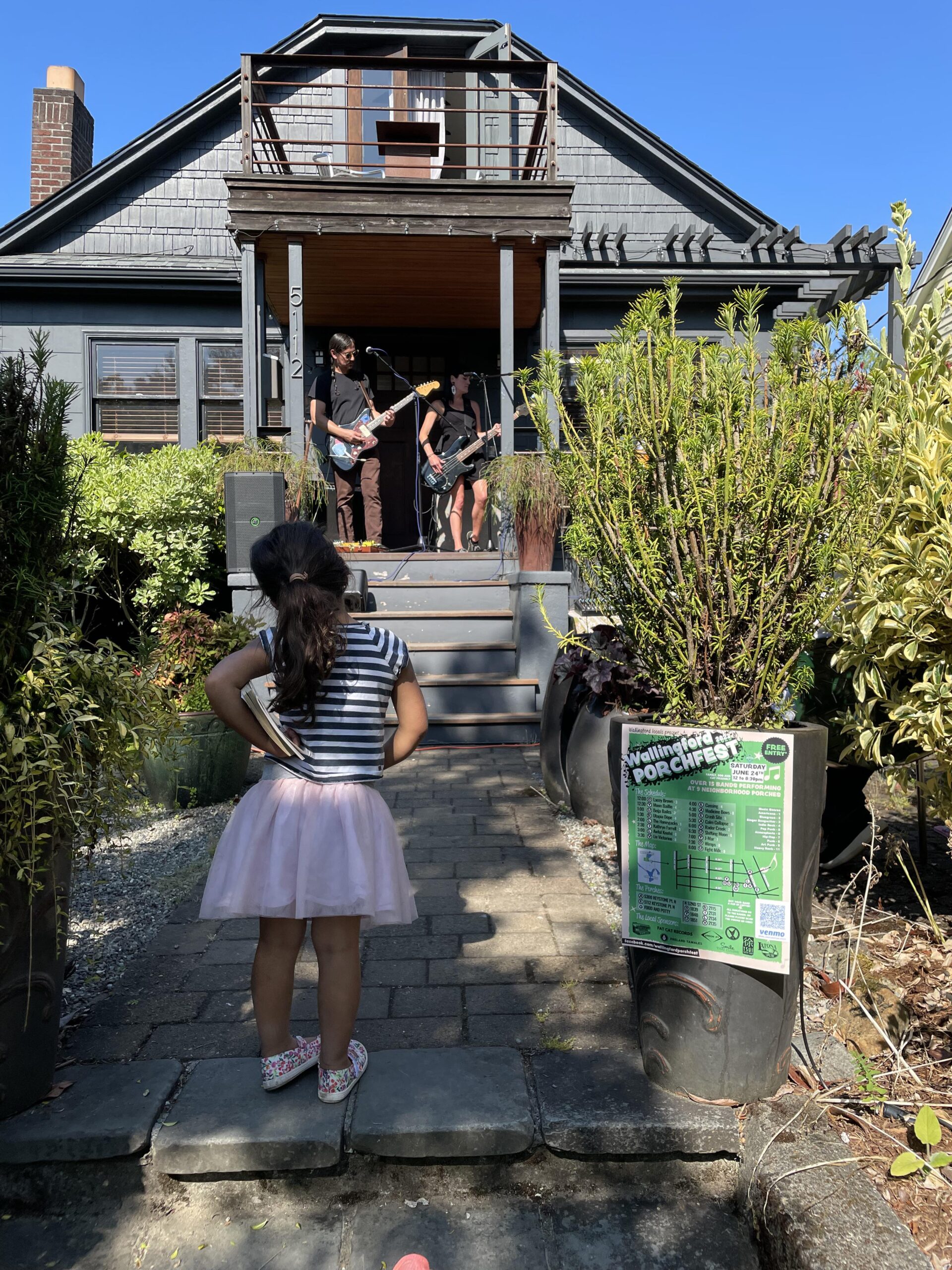 A young girl wearing a striped shirt and pink tutu watches two musicians perform on a residential front porch.