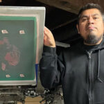 A Latino man with short black hair and a goatee standing next to a printmaking press.
