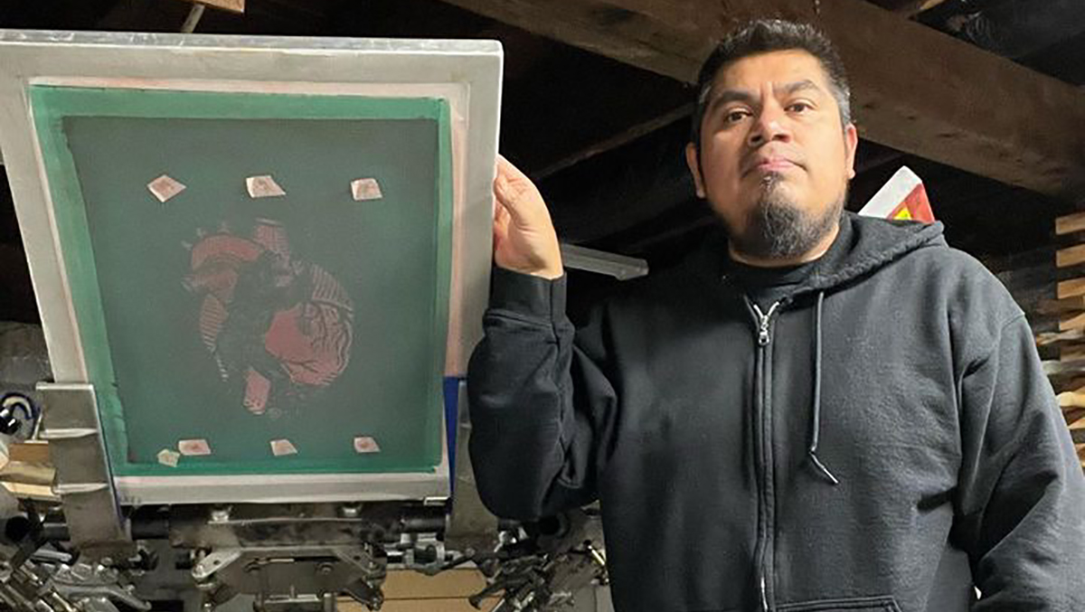 A Latino man with short black hair and a goatee standing next to a printmaking press.