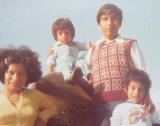 A vintage photo of a Latino family with a woman who has short brown hair wearing a yellow shirt, a man wearing a red a white sweater vest over a white button up shirt, and you children. 
