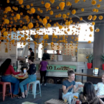 Customers sit at tables at a covered patio of a food truck with orange decoration hanging from the ceiling.
