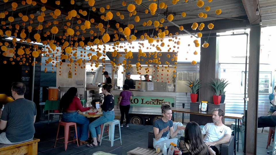 Customers sit at tables at a covered patio of a food truck with orange decoration hanging from the ceiling.