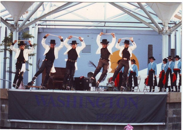 A stage with five performers dancing in black pants, shirt, and hats with white jackets.