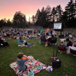 Groups of people sitting on picnic blankets in a park lawn at sunset watching an outdoor movie on a large projector screen