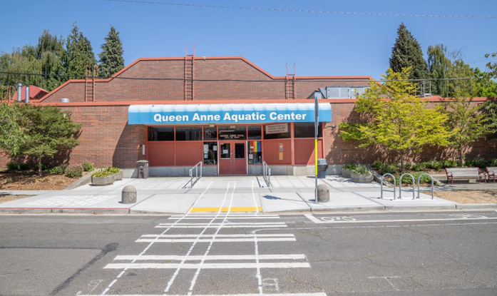 A brick, one-story building with a blue sign that has white lettering. The sign says Queen Anne Aquatic Center.