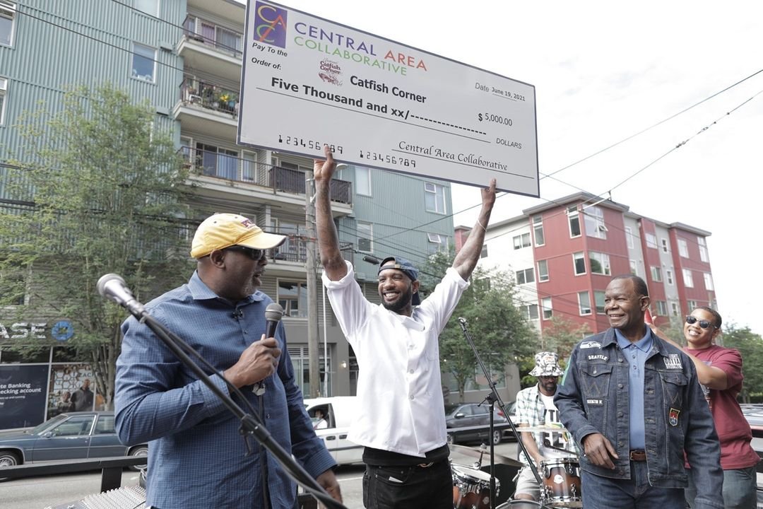 Man in white shirt holds a giant check for his business. He is smiling. Two people join him and celebrate.