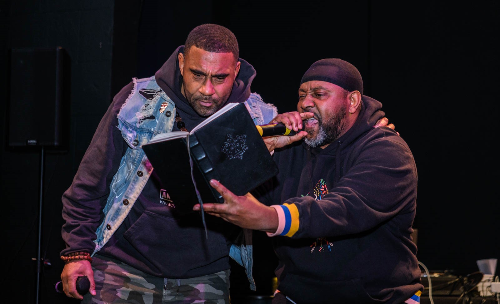 A man wearing holds a microphone to his lips as he reads from a book he is reading. Another man wearing a blue jean jacket is silently reading from the book too. 