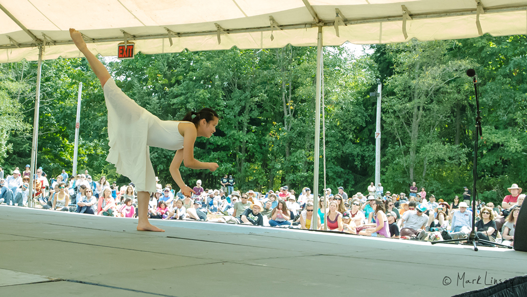 A dancer wearing a flowy white dress pictured leaning forward with one leg kicked high in the air.