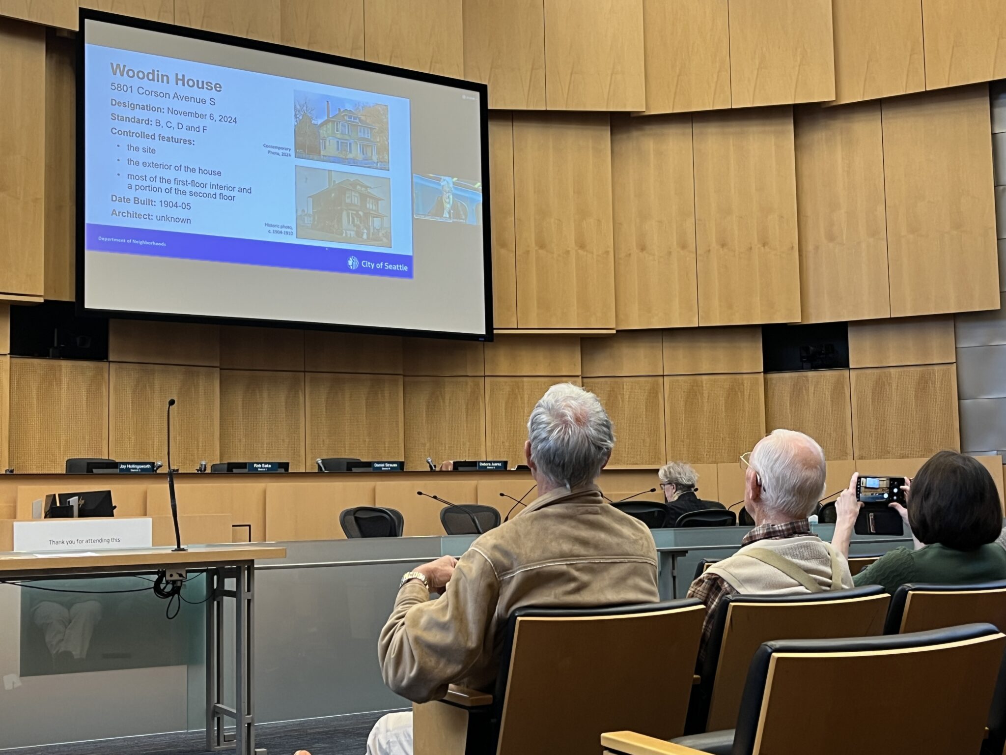 People sitting in City Council Chambers looking up at a screen with a presentation featuring a photo of a house and text that says "Woodin House"
