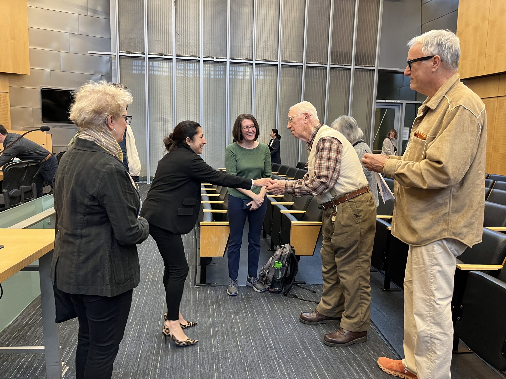 An older man wearing a sweater vest and plaid shirt shakes hands with a woman with black hair pulled back and black pants and suit jacket