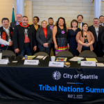 A group of eighteen people stand together behind a table with a black tablecloth with the City of Seattle logo and text that reads: "City of Seattle Tribal Nations Summit, May 2, 2023."