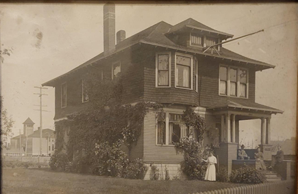 yellowed antique photo with a large house with dormers and a woman in a white dress standing in front, a little bit ghostly.