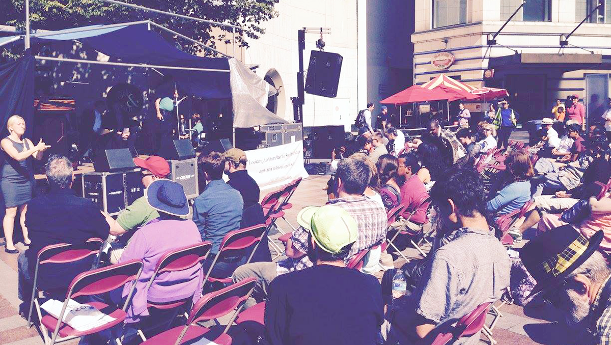 An older photograph of a crowd sitting in chairs facing a stage in a city square. A sign language interpreter signs next to the stage.