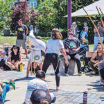 A group of people sit in a circle in a park with two people in the center doing dance and acrobatic moves