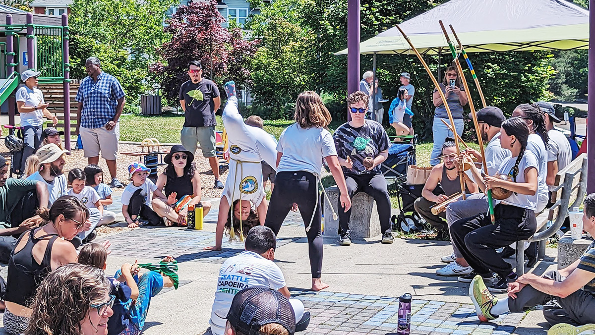 A group of people sit in a circle in a park with two people in the center doing dance and acrobatic moves