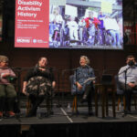 Four people sitting on a stage with microphones in front of each. From left to right, a White woman wearing a lavender sweater sitting in an electric wheelchair, a White woman wearing a black shirt and long skirt, a White woman wearing a blue dress, and a young White man wearing a light blue button up shirt and black pants. Behind them is a large screen with a photo of a group of people in wheelchairs holding picket signs and text that says "Disability Activism History"