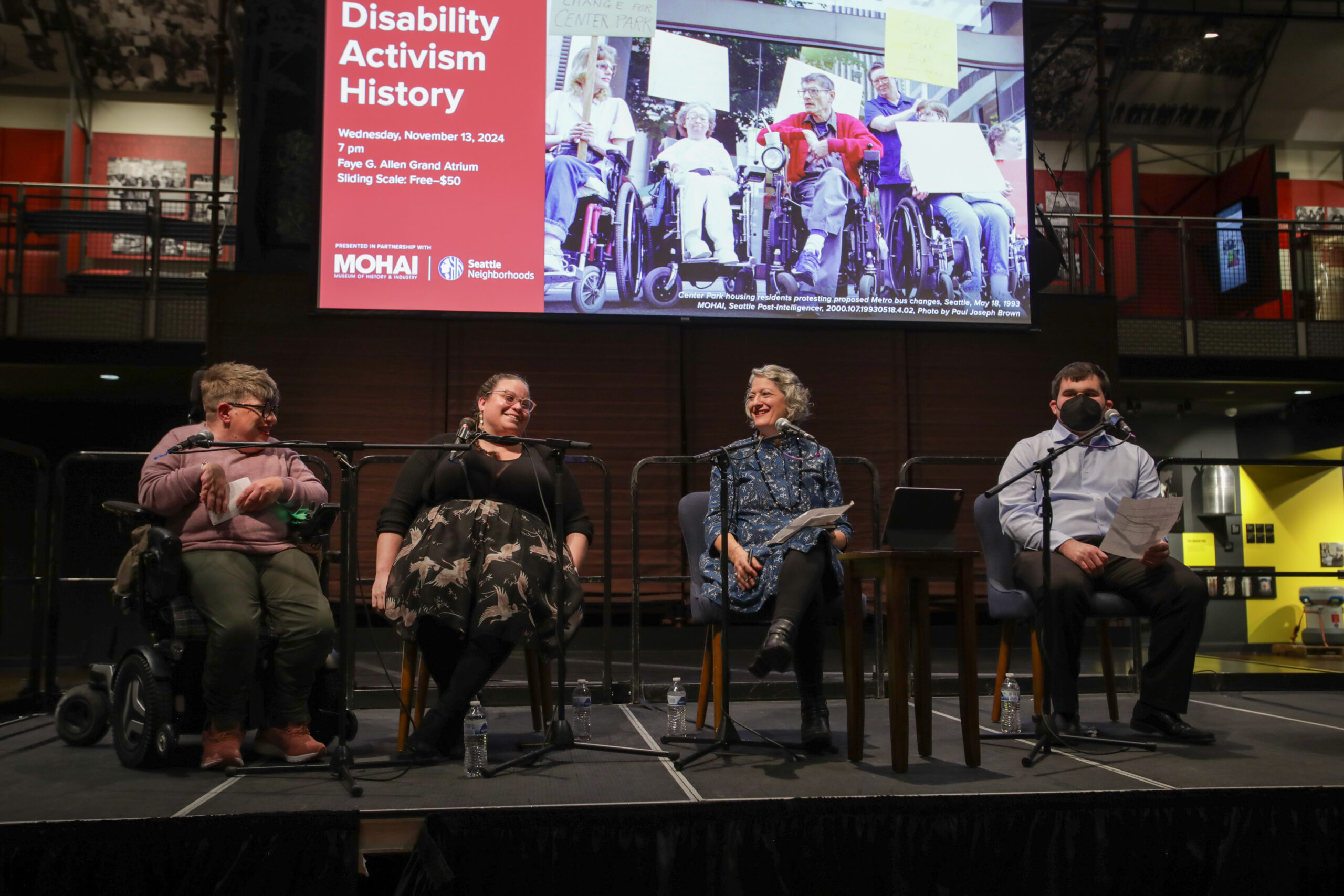 Four people sitting on a stage with microphones in front of each. From left to right, a White woman wearing a lavender sweater sitting in an electric wheelchair, a White woman wearing a black shirt and long skirt, a White woman wearing a blue dress, and a young White man wearing a light blue button up shirt and black pants. Behind them is a large screen with a photo of a group of people in wheelchairs holding picket signs and text that says 