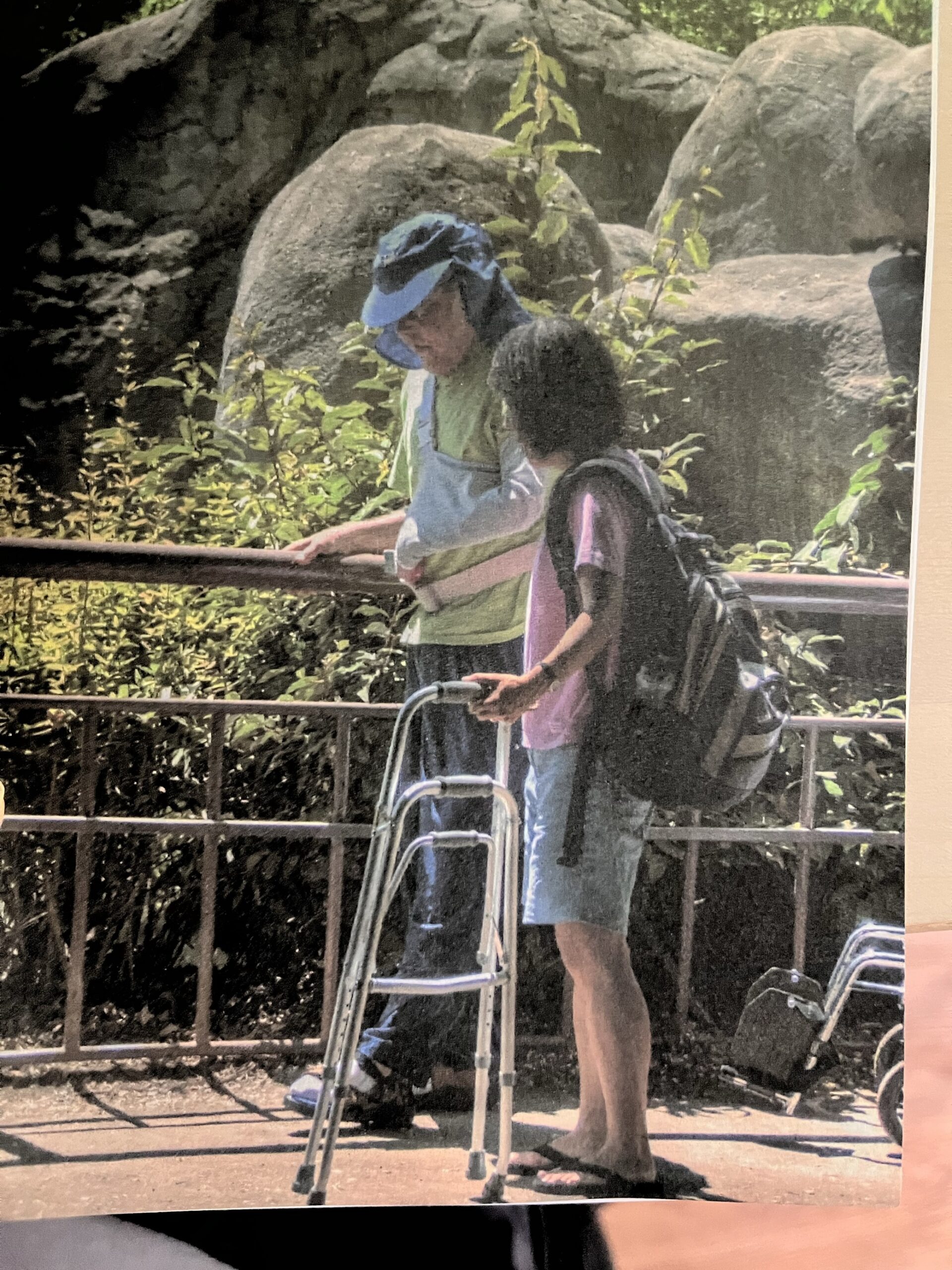 Photo of a man wearing a blue hat holding a hand rail with his right arm and his left arm in a sling. A woman to his left is helping him walk and holding a walker. Greenery and boulders are pictured in the background. 