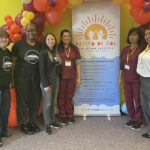 A group of people standing beneath an arch of yellow, orange, and maroon balloons with a sign in the center that reads: "Rayito De Sol Childcare Services Cooperative."