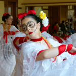 A dancer wearing a white dress with red ribbons and flowers in their hair with skeleton like paint on their face