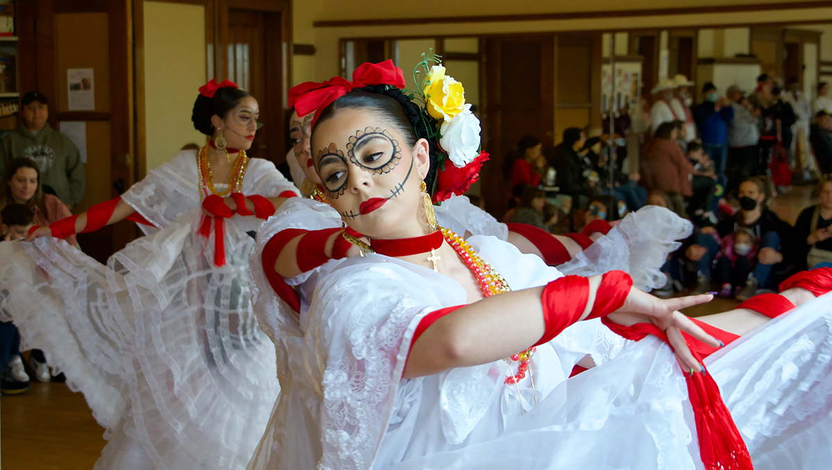 A dancer wearing a white dress with red ribbons and flowers in their hair with skeleton like paint on their face