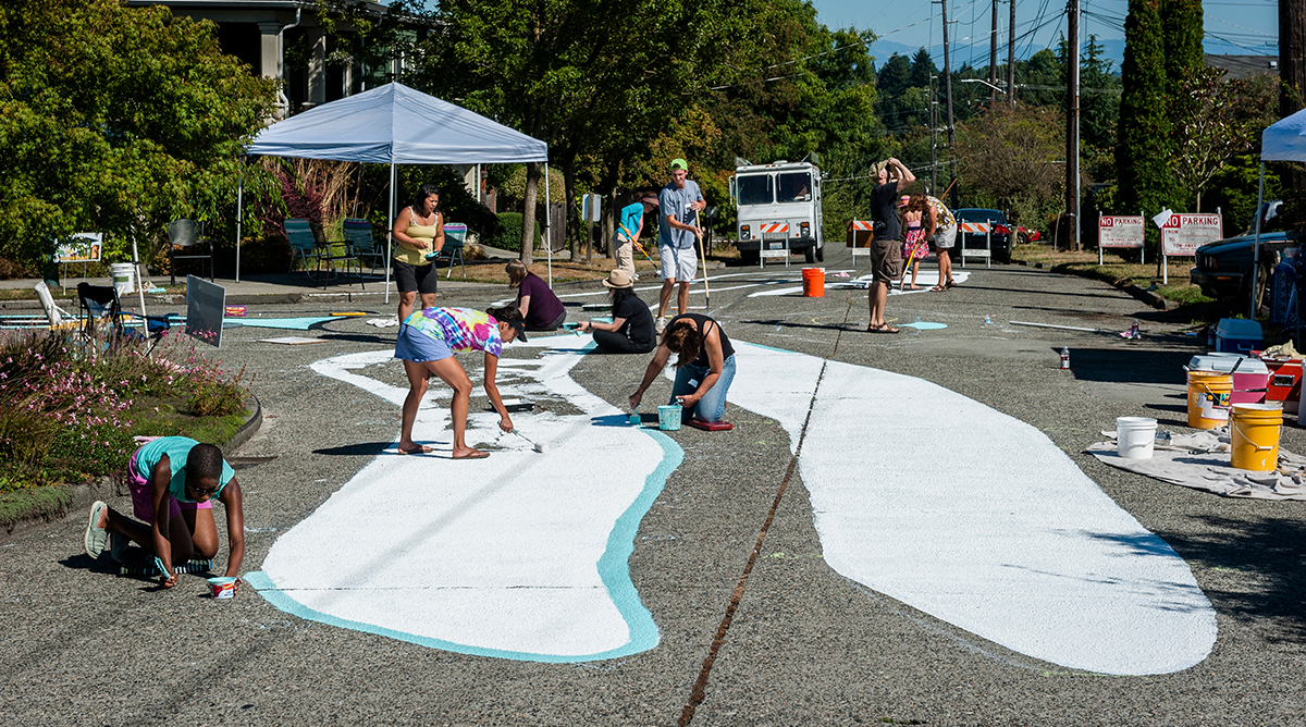 A group of people painting a large mural in the middle of a traffic intersection.