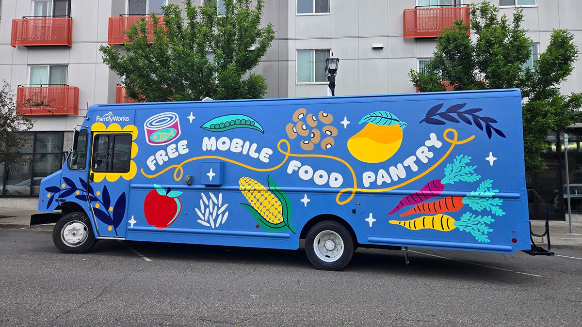 A blue box truck parked on a street next to an apartment building with bright images of food painted on the side and text that reads: 
