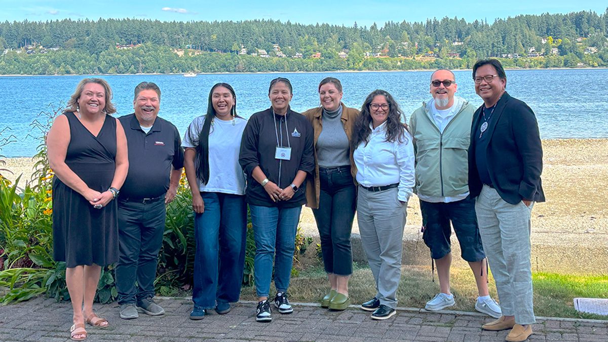 A group of eight people standing in a line smiling at the camera with a body of water and evergreen trees in the background
