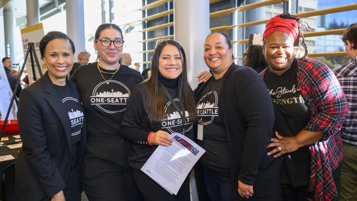 Five people stand together, smiling, wearing black shirts that say