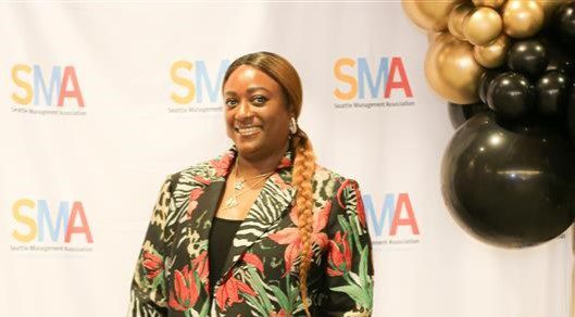 A woman with dark skin and long brown hair wearing a floral jacket smiling in front of 