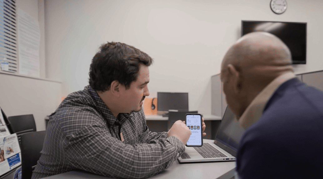 Two people sitting on at a desk with a lab top computer and looking at a mobile phone together