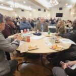A group of people sitting at a round table having a conversation. Additional tables of people are pictured in the background.