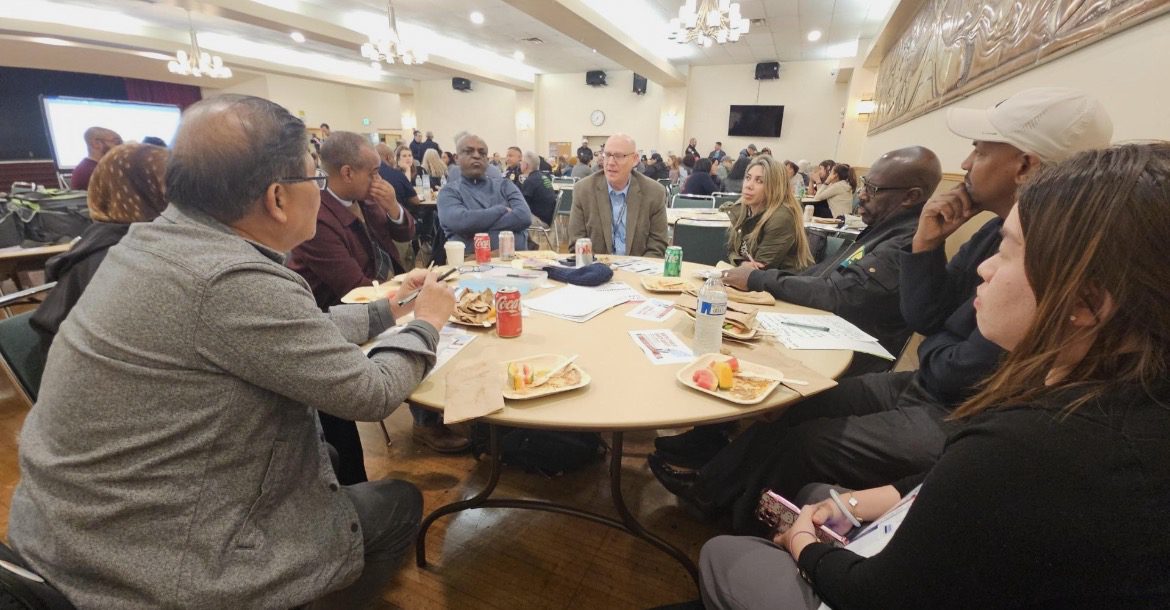 A group of people sitting at a round table having a conversation. Additional tables of people are pictured in the background.