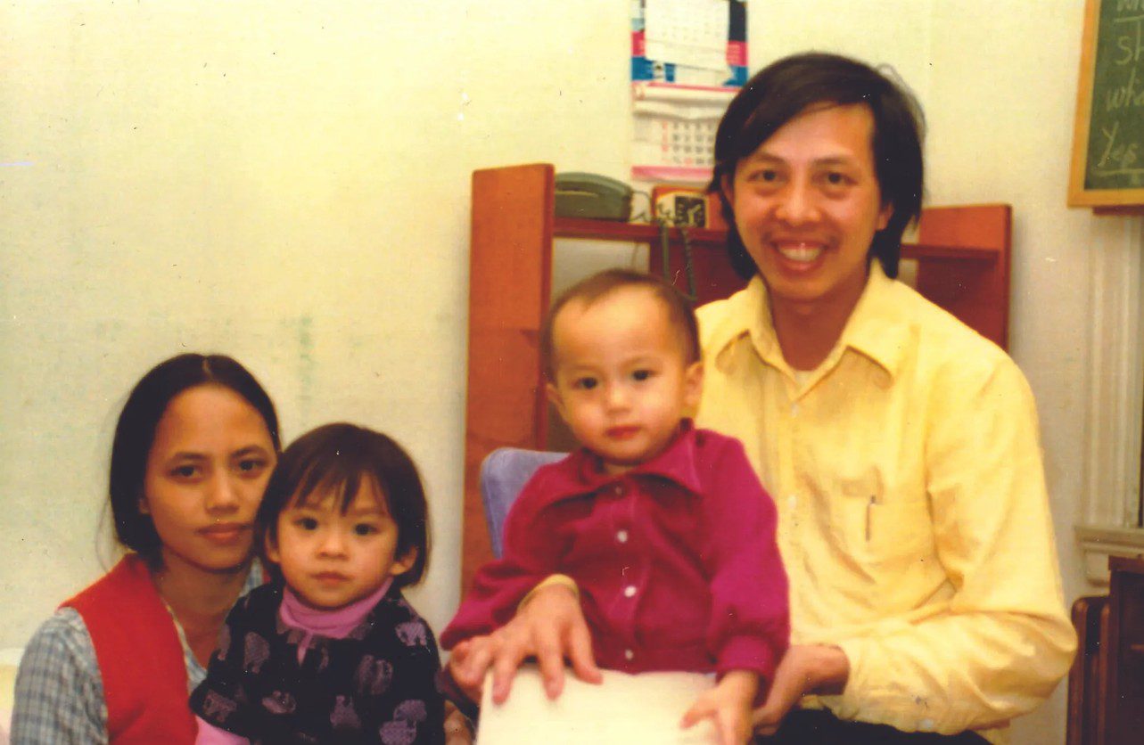 A young woman with dark black hair sitting, holding a toddler with short black hair on her lap next to a man with short black hair sitting higher up with a younger child on his lap