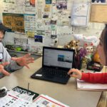 An older person sits across a table from a younger person as they look at a laptop computer