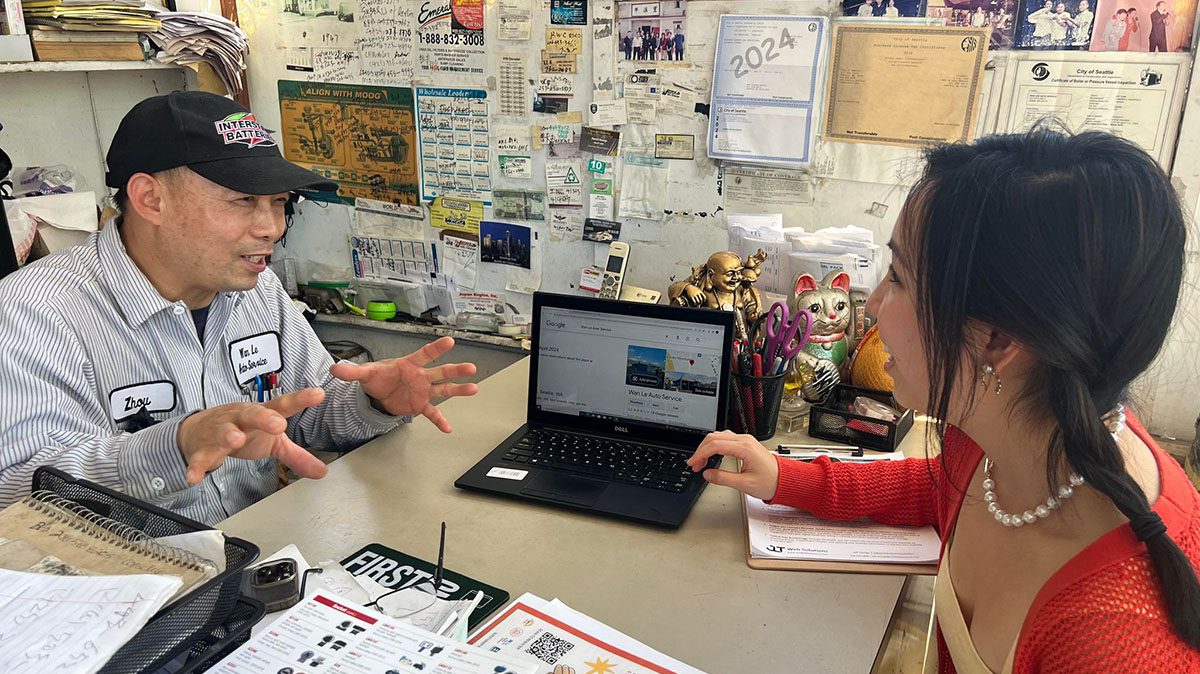 An older person sits across a table from a younger person as they look at a laptop computer