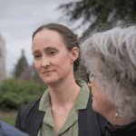 A white female with light brown hair pulled back, wearing a green collard shirt listening pensively to two people with gray hair in the foreground.