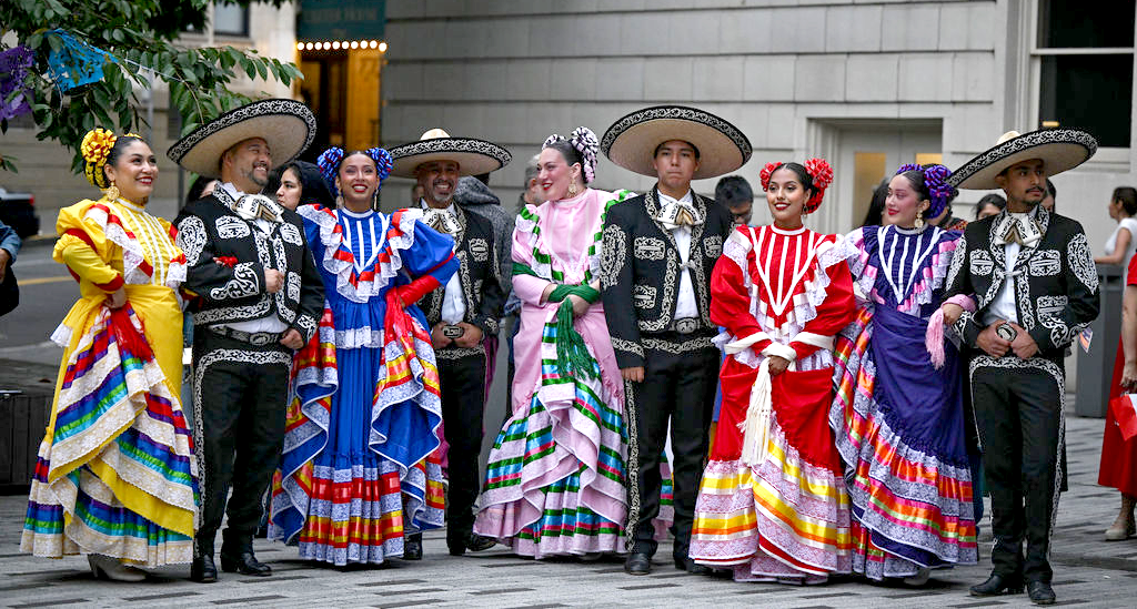 A group of dancers in traditional Mexican attire standing in a line in front of a gray brick building
